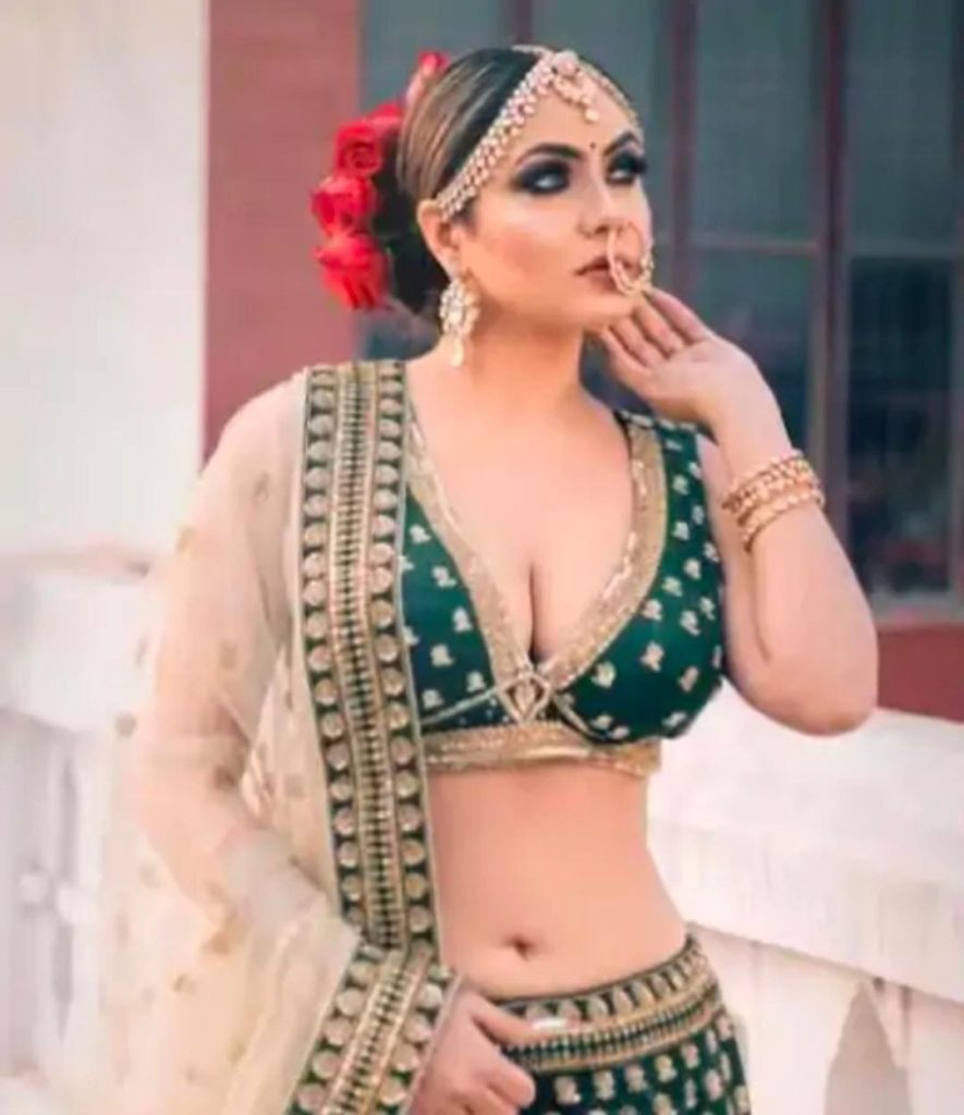 Woman in a green heavily embellished lehenga with gold jewelry, red flowers in her bun, posing with her hand near her face.