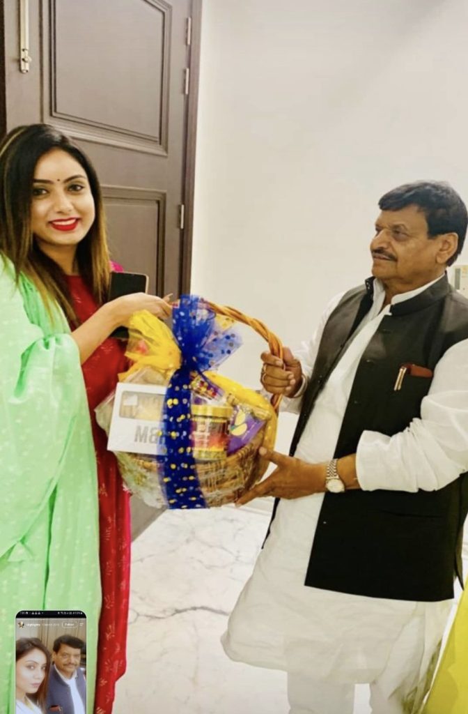 Woman in a red sari and green shawl offers a gift basket to a man in white kurta and black vest; basket wrapped in blue and yellow ribbons.