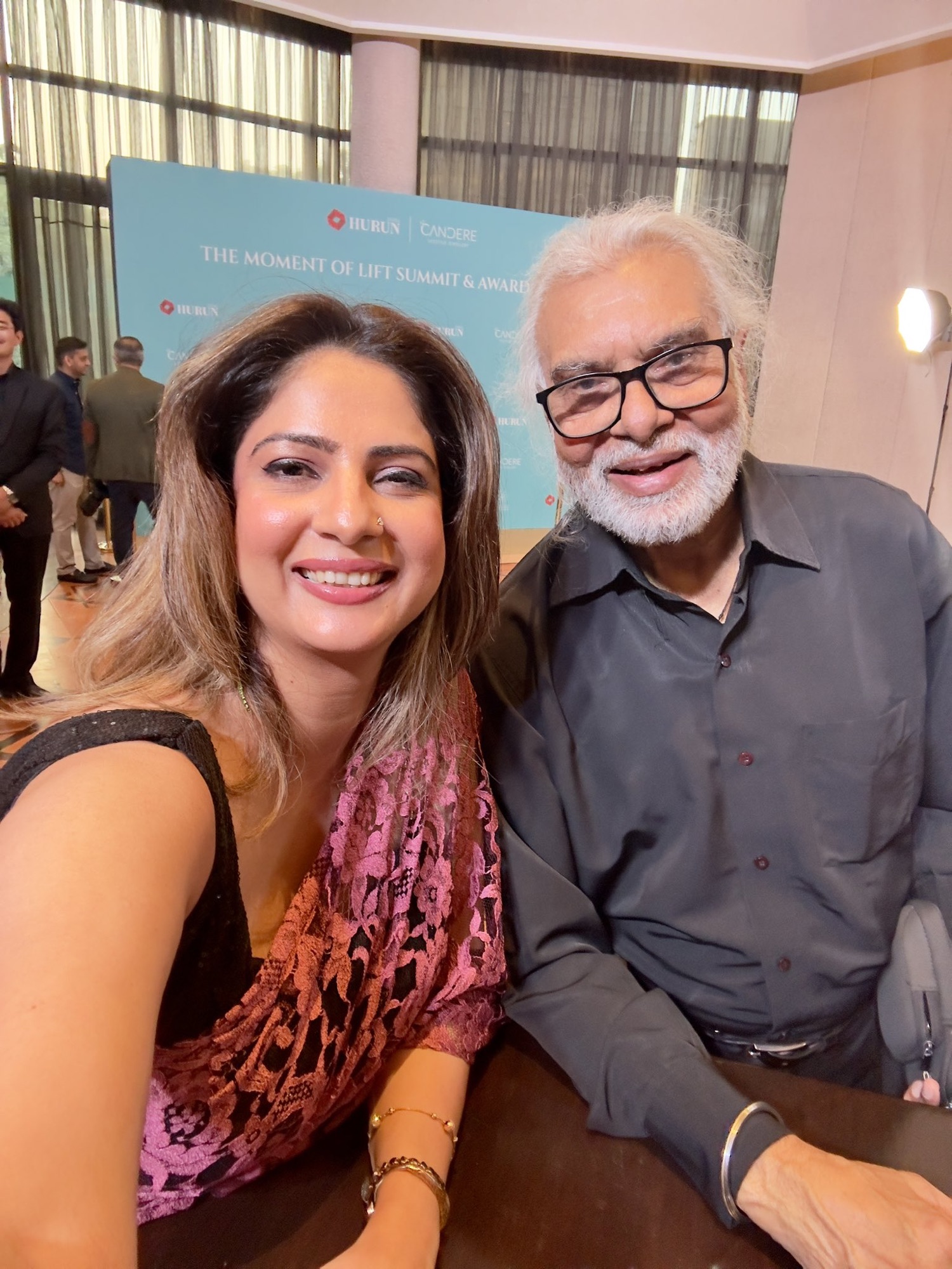 Two people smiling for a selfie at an event, with a blue backdrop reading 'The Moment of Lift Summit & Awards'.