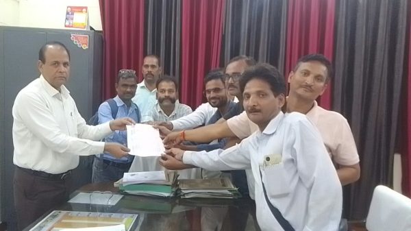 A group of men in an office passing a document to each other over a cluttered desk during a meeting or ceremony.