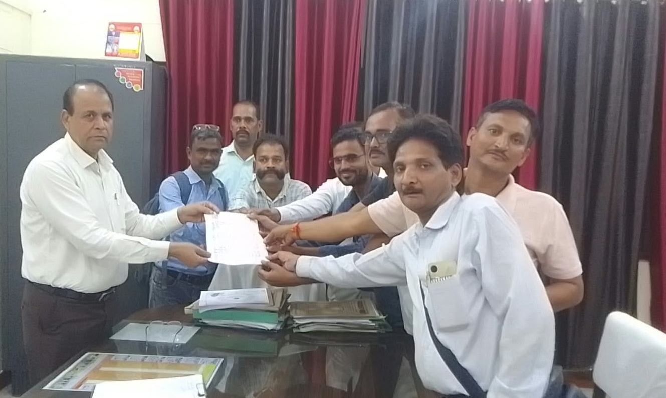 A group of men in an office passing a document to each other over a cluttered desk during a meeting or ceremony.