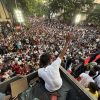 A man in a white shirt sits on a platform at the edge of a massive crowd gathered for a street rally, raising his hand to address the protesters.