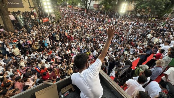 A man in a white shirt sits on a platform at the edge of a massive crowd gathered for a street rally, raising his hand to address the protesters.