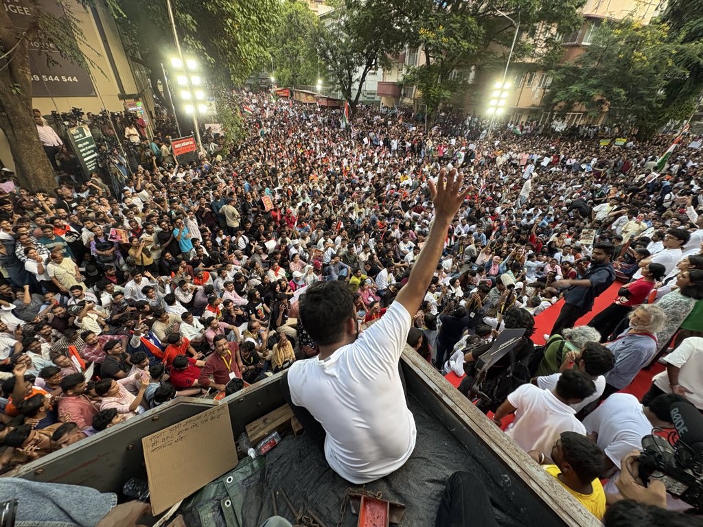 A man in a white shirt sits on a platform at the edge of a massive crowd gathered for a street rally, raising his hand to address the protesters.
