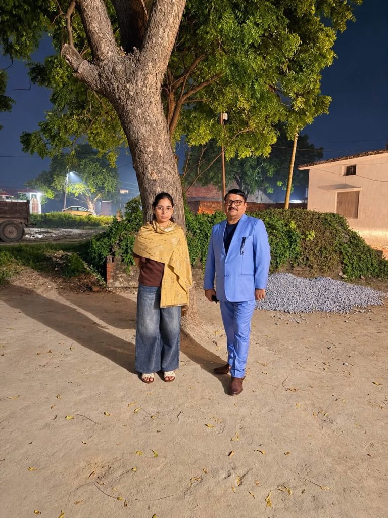 Two people pose under a large tree at night: a woman in a tan scarf and jeans, and a man in a blue suit.