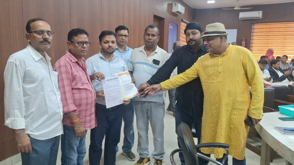 Seven men in a wood-paneled office exchange a document; a man in a yellow kurta and hat extends his hand toward the papers as others look on.