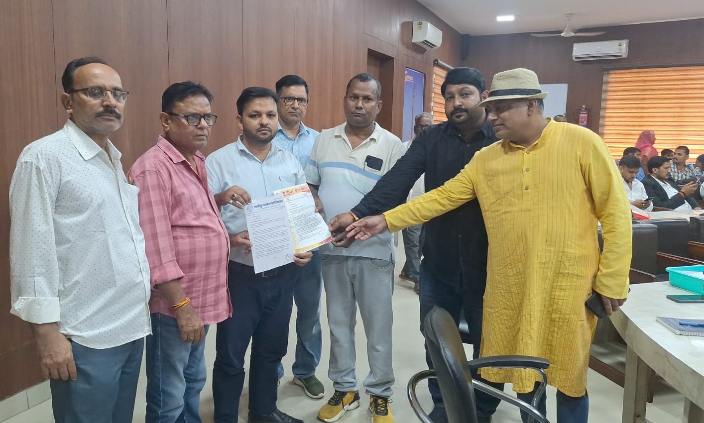 Seven men in a wood-paneled office exchange a document; a man in a yellow kurta and hat extends his hand toward the papers as others look on.