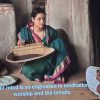 Woman in a teal saree with pink border sits cross‑legged holding a woven basket, inside a rustic room with pottery nearby.