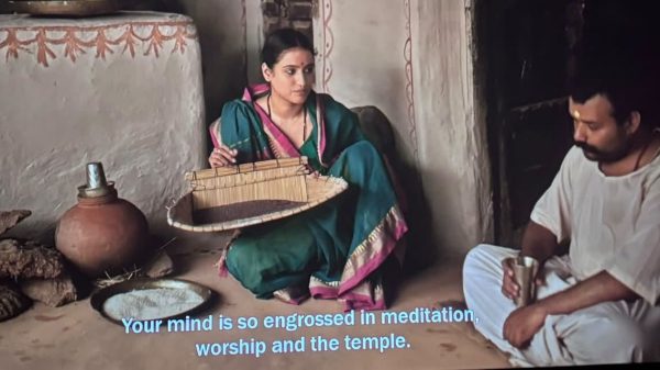 Woman in a teal saree with pink border sits cross‑legged holding a woven basket, inside a rustic room with pottery nearby.