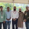 Group of eight men standing side by side under a shaded pavilion outdoors, posing for a group photo, some wearing lanyards and casual attire.