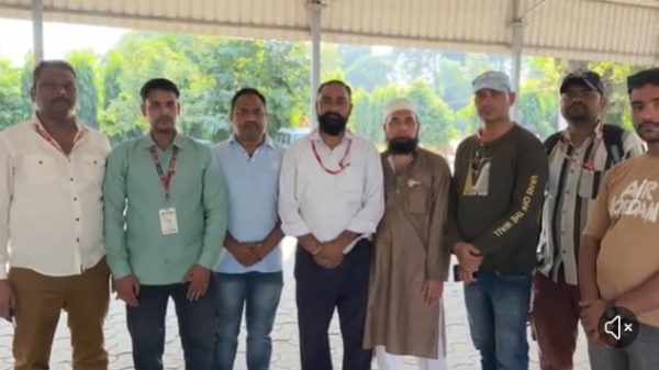 Group of eight men standing side by side under a shaded pavilion outdoors, posing for a group photo, some wearing lanyards and casual attire.