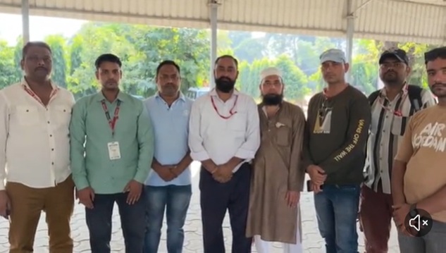 Group of eight men standing side by side under a shaded pavilion outdoors, posing for a group photo, some wearing lanyards and casual attire.