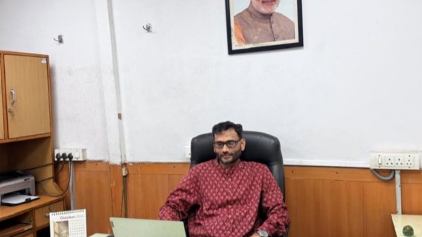 Man in a red patterned shirt seated at a desk with a laptop in an office; a framed portrait hangs on the wall behind him.
