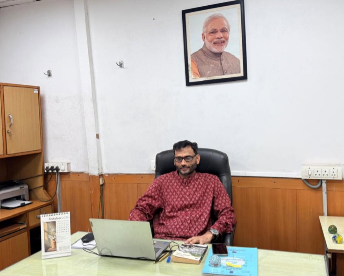 Man in a red patterned shirt seated at a desk with a laptop in an office; a framed portrait hangs on the wall behind him.