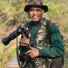 Photographer standing outdoors on a wooden platform, wearing a camo vest and hat, holding a DSLR with a large telephoto lens.