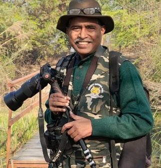 Photographer standing outdoors on a wooden platform, wearing a camo vest and hat, holding a DSLR with a large telephoto lens.