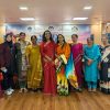 Ten women in colorful sarees and outfits stand together on a stage for a group photo at a conference or event with a banner behind them.