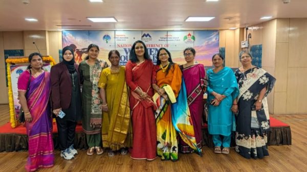 Ten women in colorful sarees and outfits stand together on a stage for a group photo at a conference or event with a banner behind them.