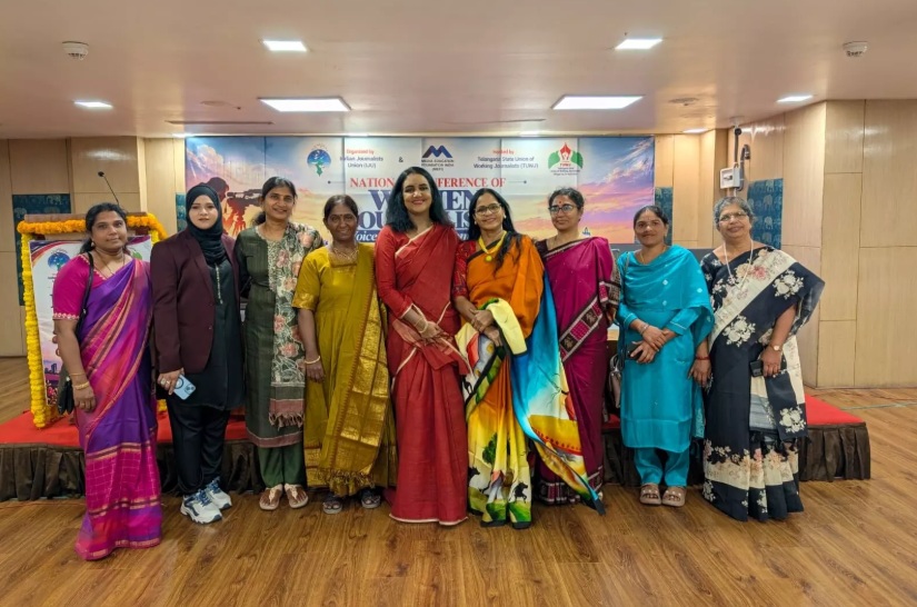 Ten women in colorful sarees and outfits stand together on a stage for a group photo at a conference or event with a banner behind them.