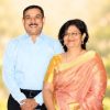 A smiling couple stands outdoors on a sunny day; the man wears a light blue shirt and dark trousers, the woman wears a peach saree with a gold border and gold jewelry.