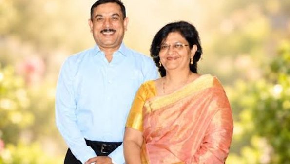 A smiling couple stands outdoors on a sunny day; the man wears a light blue shirt and dark trousers, the woman wears a peach saree with a gold border and gold jewelry.