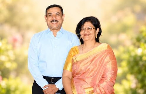 A smiling couple stands outdoors on a sunny day; the man wears a light blue shirt and dark trousers, the woman wears a peach saree with a gold border and gold jewelry.
