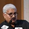 Man with white hair and glasses speaks into microphones at a podium.