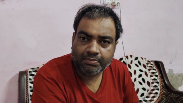 Man with short dark hair and beard wearing a red shirt sits on a couch indoors, looking at the camera.