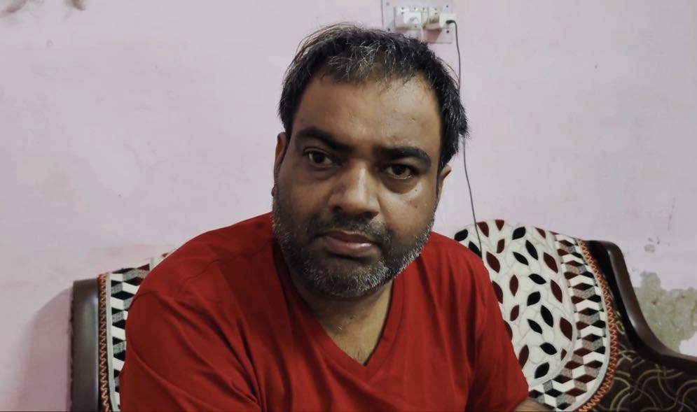 Man with short dark hair and beard wearing a red shirt sits on a couch indoors, looking at the camera.