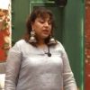 Woman wearing a light gray traditional kurta with large silver earrings, standing indoors and speaking to an audience.