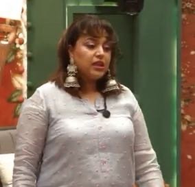 Woman wearing a light gray traditional kurta with large silver earrings, standing indoors and speaking to an audience.