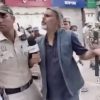 Reporter with a microphone confronts a man in a blue vest who gestures with his arm as police stand nearby in a crowded street, storefronts in the background.