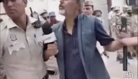 Reporter with a microphone confronts a man in a blue vest who gestures with his arm as police stand nearby in a crowded street, storefronts in the background.