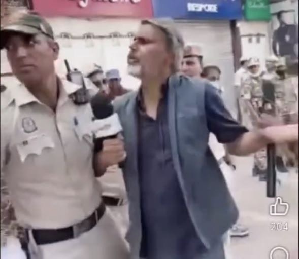 Reporter with a microphone confronts a man in a blue vest who gestures with his arm as police stand nearby in a crowded street, storefronts in the background.