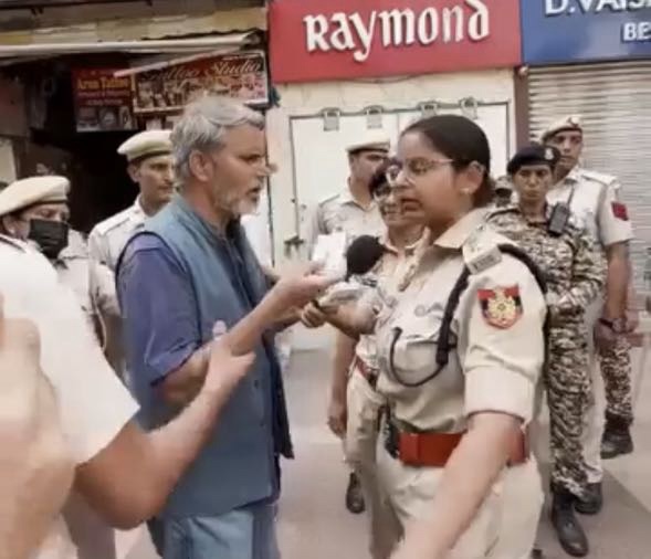 Bearded man in a blue vest being interviewed by a female police officer in khaki uniform on a street, with other officers in the background.