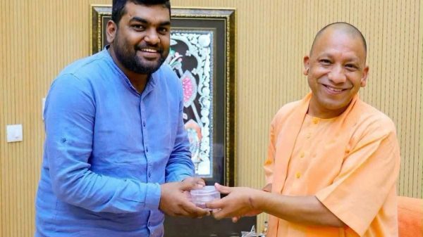 Two men exchange a small container indoors, both smiling at the camera.