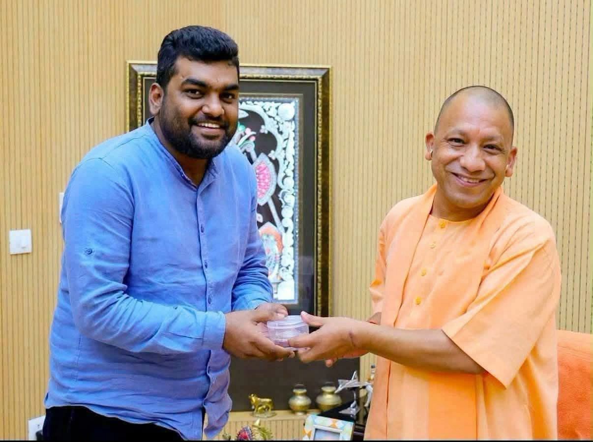 Two men exchange a small container indoors, both smiling at the camera.