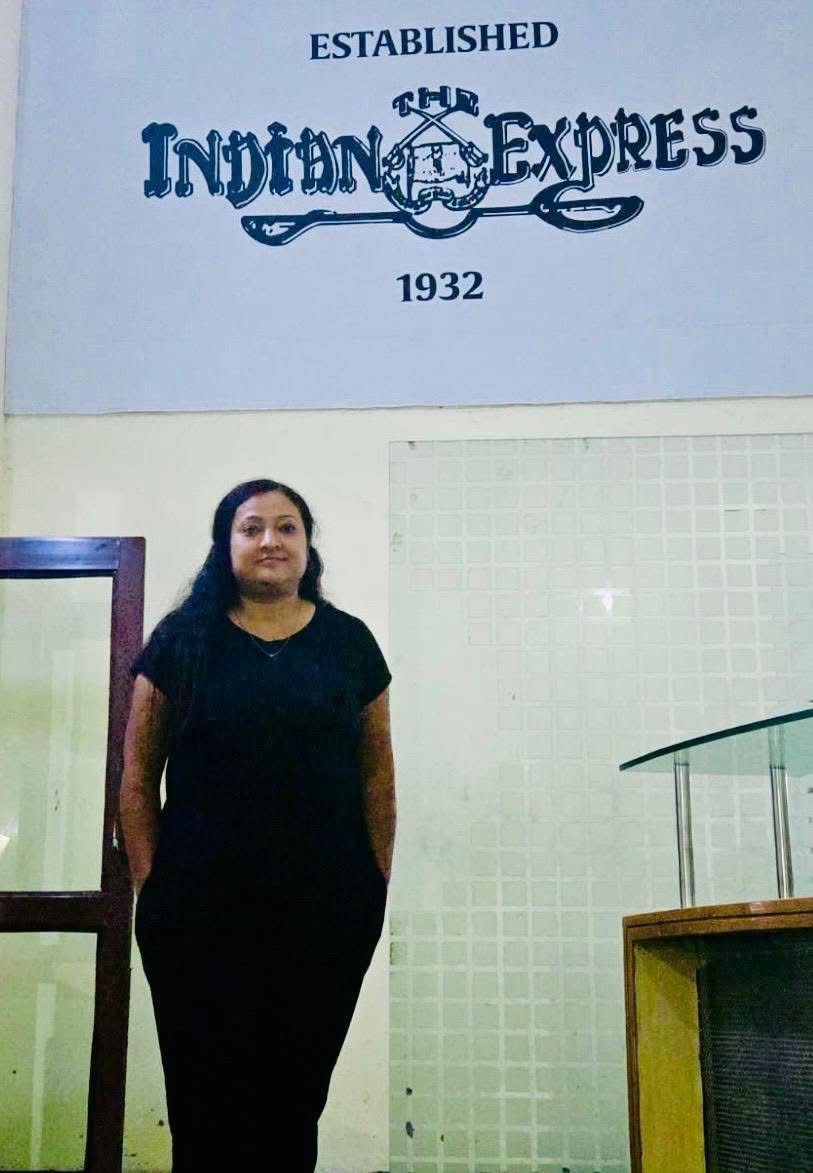 Woman in a black dress standing with hands at her sides in front of a wall sign reading 'Established Indian Express 1932'.