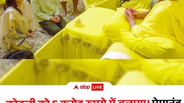 Group of people in yellow robes and light clothing sit and kneel around a yellow-clad speaker at a ceremonial or religious gathering, with a microphone on the table and a banner-like background behind them.