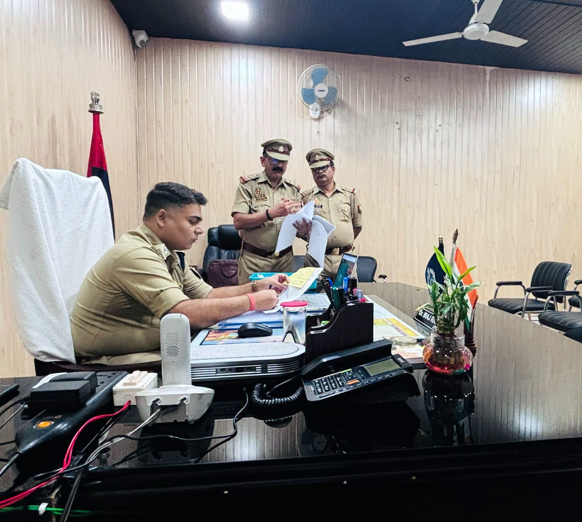 Police officers in khaki uniforms review documents at a conference table in an office room.
