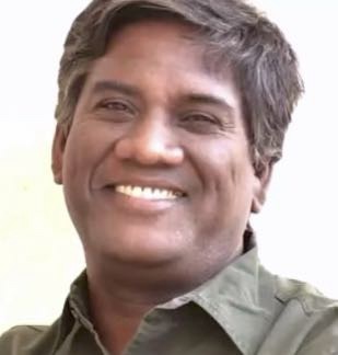 Screenshot | No. 1 Indian Media News Portal Smiling man with short dark hair wearing a green collared shirt, facing the camera against a light background.