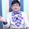 Presenter in glasses wearing a light blazer and purple zigzag scarf speaks to the camera in a studio with blue vertical stripes, gesturing with one hand on a desk.