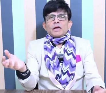 Presenter in glasses wearing a light blazer and purple zigzag scarf speaks to the camera in a studio with blue vertical stripes, gesturing with one hand on a desk.