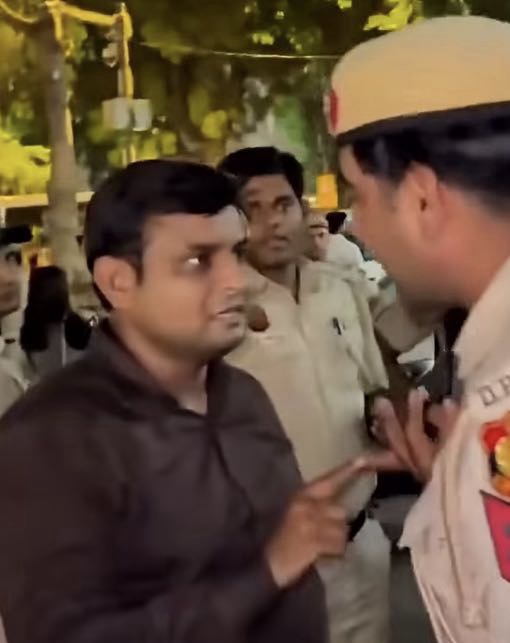 Man in a dark shirt points his finger at a uniformed officer amid a crowd outdoors, signaling a confrontation.