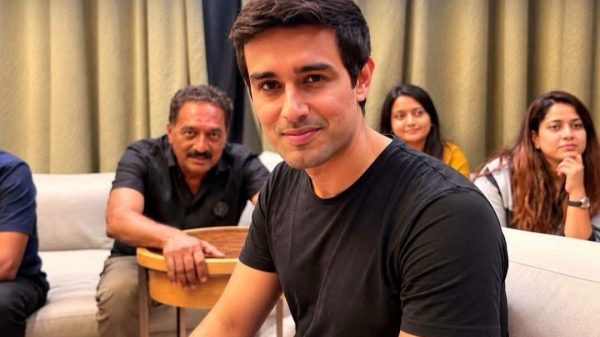A young man in a black t-shirt sitting on a sofa, with three people in the background smiling and watching.