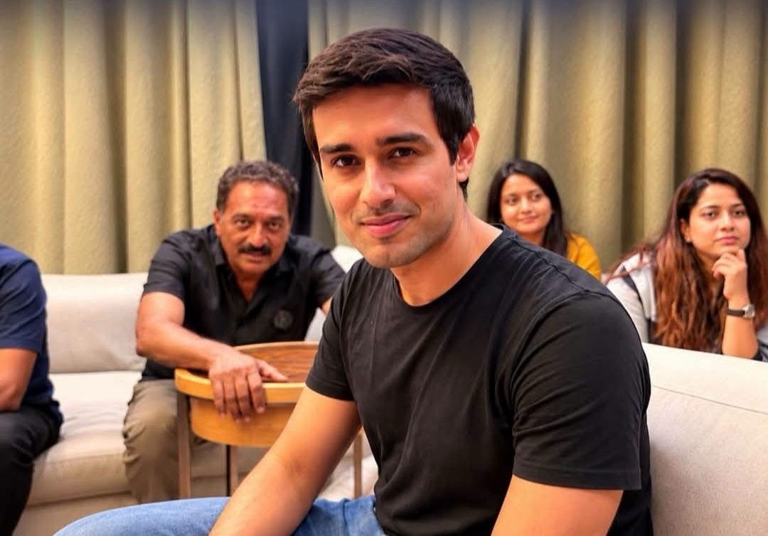 img_1905 | No. 1 Indian Media News Portal A young man in a black t-shirt sitting on a sofa, with three people in the background smiling and watching.