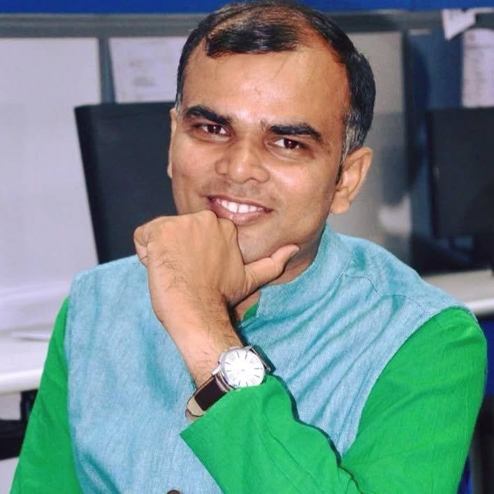 Smiling man resting his chin on his hand in an office, wearing a green kurta with a light-blue vest and a wristwatch.