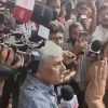 Man with white hair in a blue striped shirt speaks to a crowd of reporters; microphones, cameras, and people surround him as they record the moment.