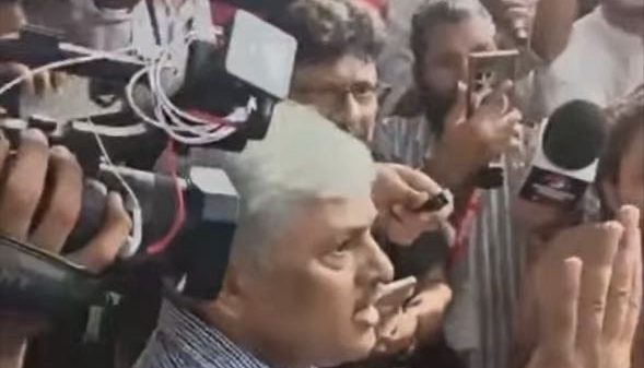 Man with white hair in a blue striped shirt speaks to a crowd of reporters; microphones, cameras, and people surround him as they record the moment.