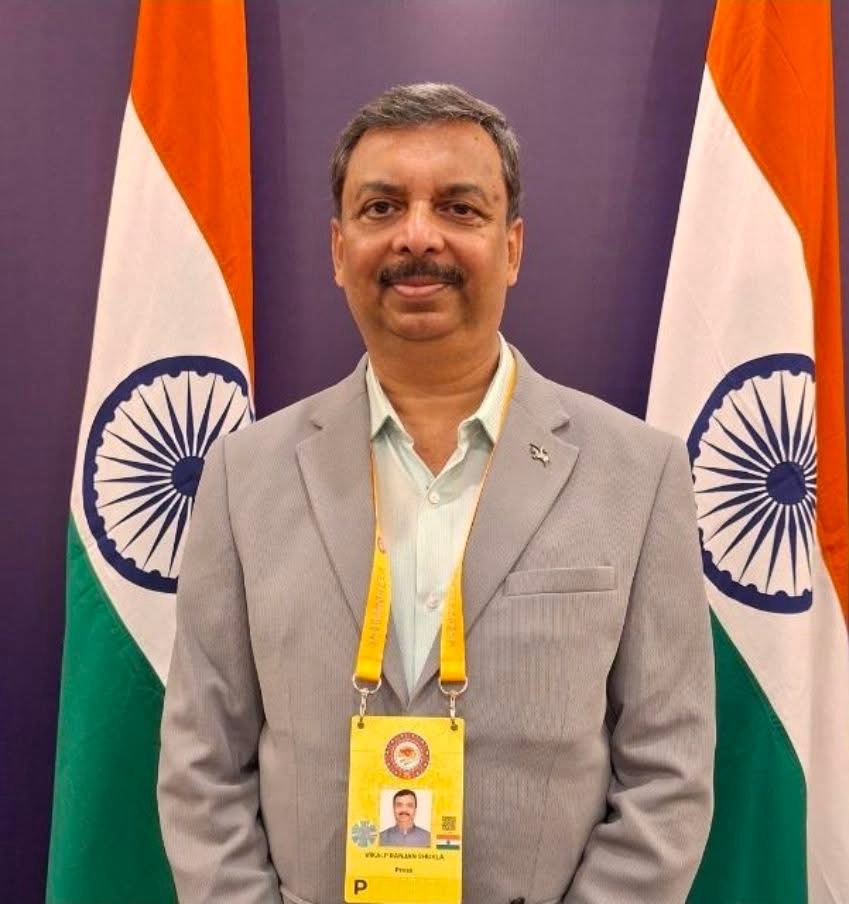 Indian man in a gray suit with a yellow lanyard and ID badge, standing in front of Indian flags and smiling.
